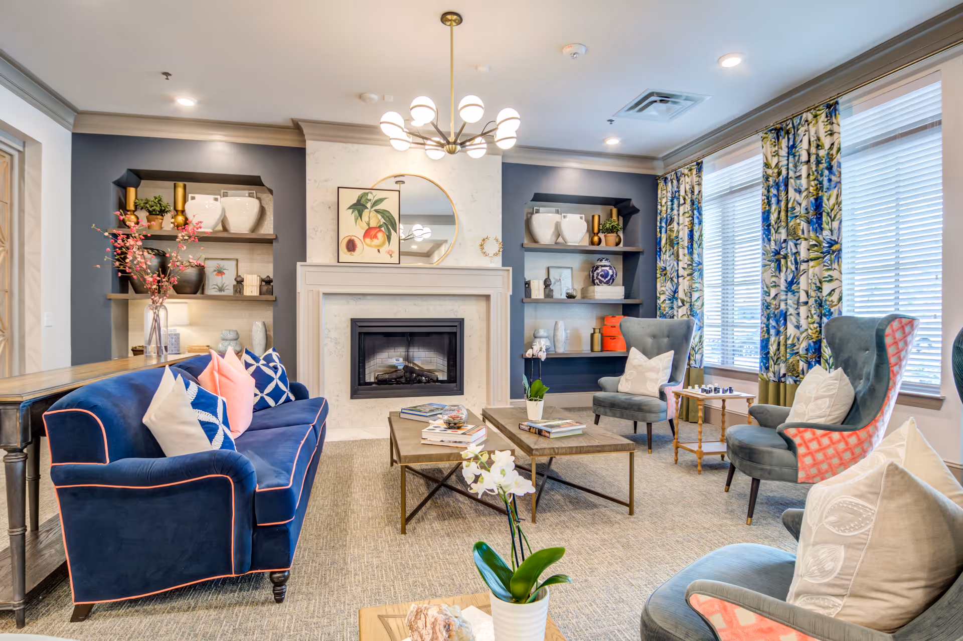 A bright and cozy living room with a blue velvet sofa adorned with colorful pillows, two gray armchairs with patterned backs, a wooden coffee table with books and a small plant, a fireplace with a round mirror and artwork above it, built-in shelves with decorative items, and large windows with floral curtains letting in natural light.