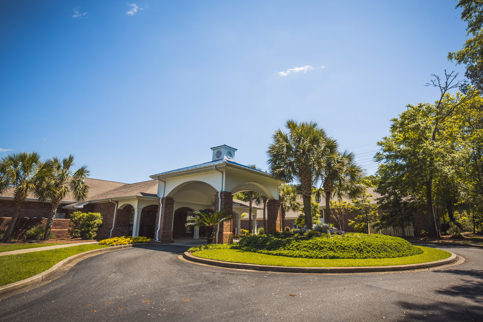 Front entrance of a brick senior living building with a covered porte-cochère, palm trees, and a circular driveway.