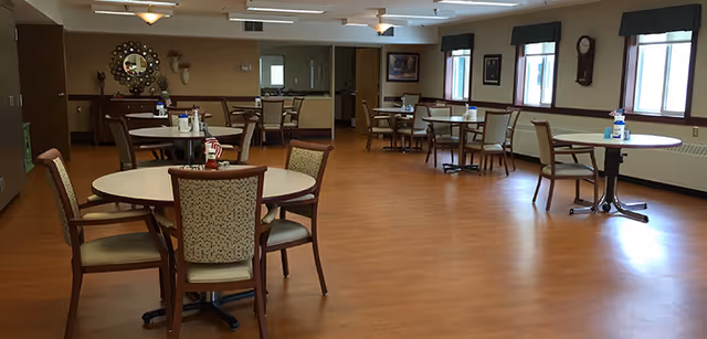 A spacious dining room with several round tables and chairs arranged neatly. The room has wooden flooring, windows with green valances, and wall decorations including a clock and framed pictures. The lighting is soft and the room appears clean and well-maintained.