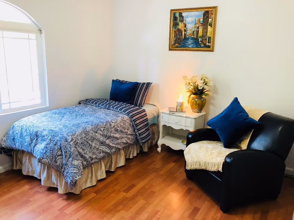 A cozy bedroom with a single bed covered in blue patterned bedding and a blue pillow. Next to the bed is a white nightstand with a lit candle, a book, and a vase of flowers. A black armchair with a blue pillow and a beige throw blanket is positioned nearby. A framed painting of a canal scene hangs on the white wall above the nightstand. The room has wooden flooring and a window with white blinds.