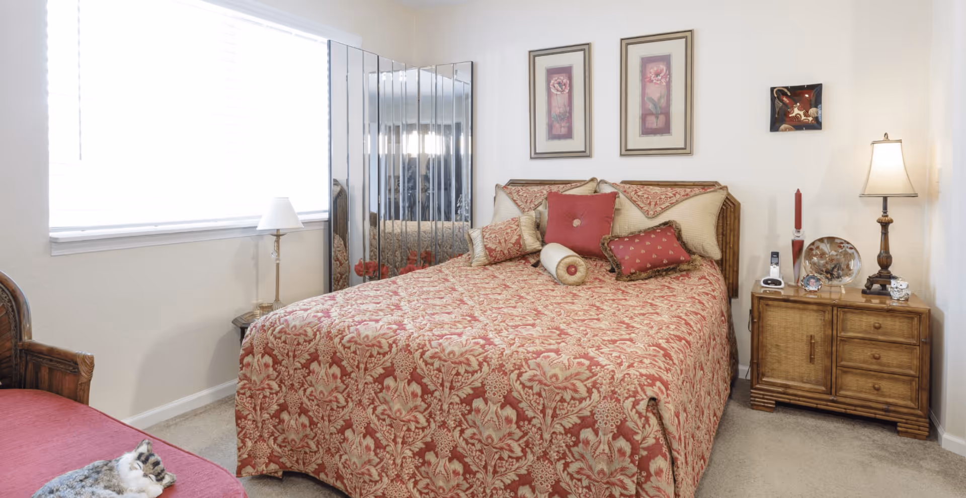 Well-lit bedroom with a patterned red bedspread, decorative pillows, wooden nightstands and lamps, and a mirrored panel by the window.