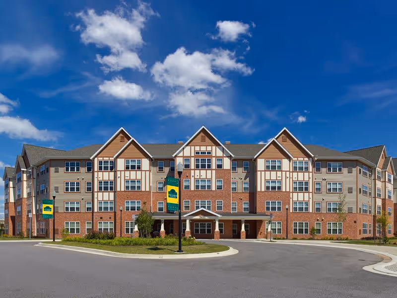 Front exterior of a four-story brick-and-siding senior living building with a central entrance and circular driveway under a blue sky.