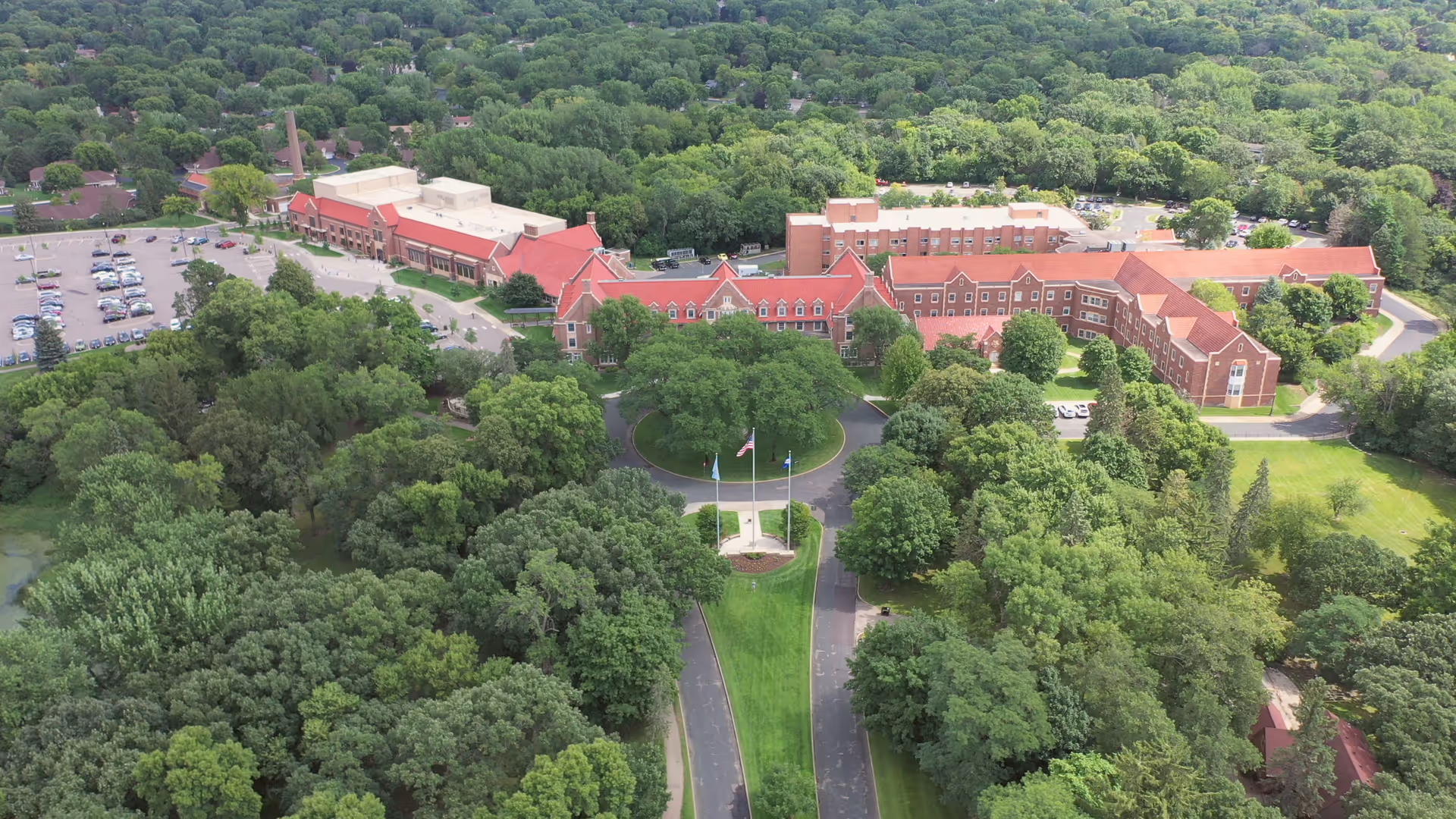 Aerial view of the Minnesota Masonic Home facility surrounded by dense green trees and landscaped lawns. The large brick building complex features red roofs and multiple wings, with a circular driveway and flagpoles at the front entrance. A parking lot with numerous cars is visible to the left, and the area is set within a lush, wooded environment.