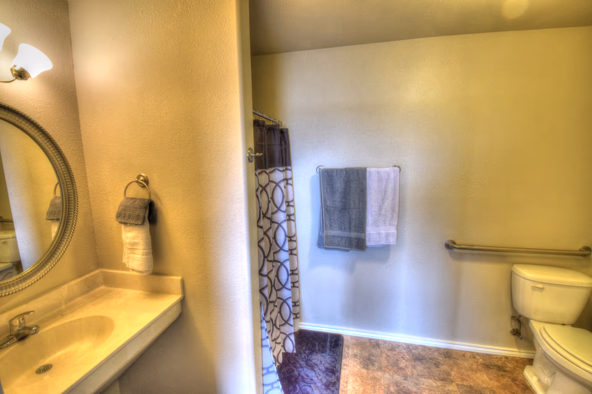 Bathroom with a sink and round mirror on the left, a shower with patterned curtain in the center, and a toilet with a grab bar on the right.