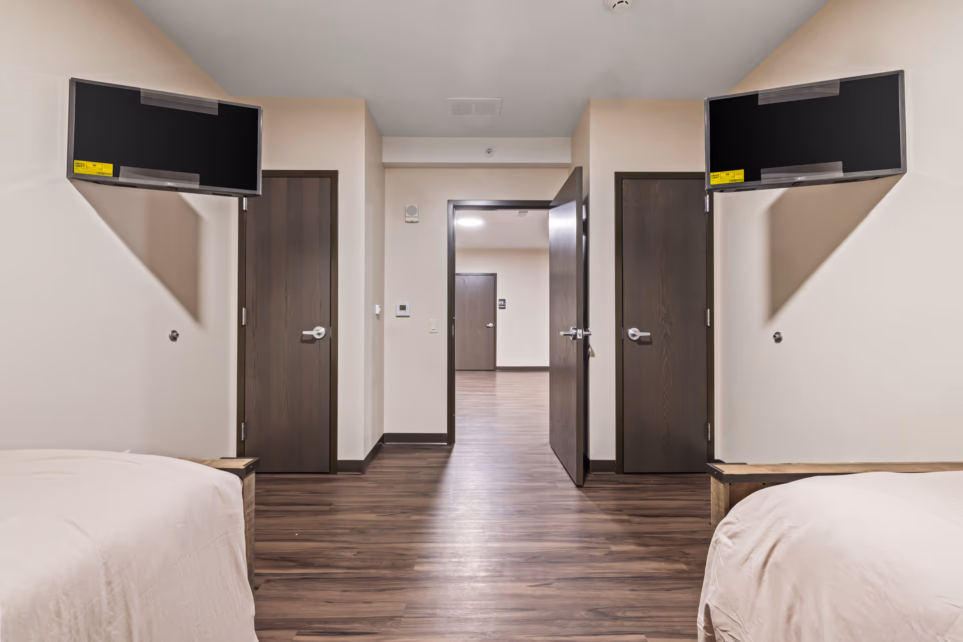 Interior view of a shared assisted-living bedroom with two beds, wall-mounted TVs, and a central doorway opening to a hallway.