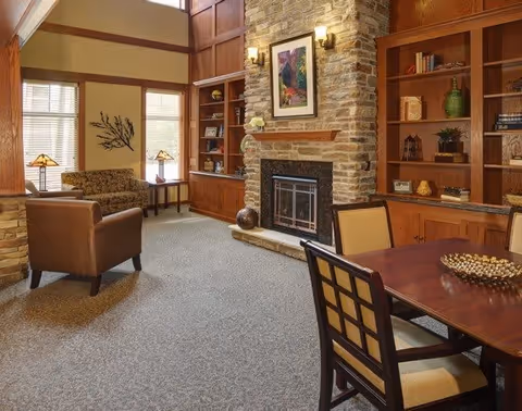 A cozy senior living common area with a stone fireplace, wooden built-in bookshelves, a table with chairs, and comfortable seating including an armchair and a loveseat. The room has large windows letting in natural light and warm wall sconces above the fireplace.