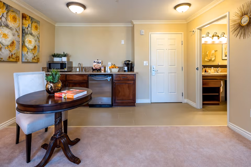 Interior view of a senior living facility room featuring a small kitchenette with wooden cabinets, a mini refrigerator, microwave, coffee maker, and a sink. In the foreground, there is a round wooden table with a white cushioned chair, a small plant in a gold vase, and two books on the table. The room has beige walls and carpeted flooring. To the right, there is an open door leading to a bathroom with a sink and mirror visible. Decorative wall art and a sunburst wall clock are also present.