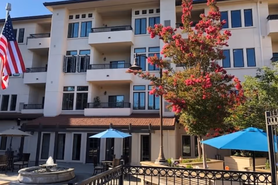 Courtyard in front of a multi-story senior living building with balconies, a fountain, blue umbrellas, and a blooming tree.