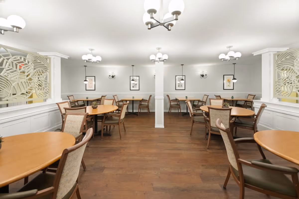 A bright and clean dining room with multiple round and rectangular wooden tables surrounded by cushioned chairs. The room features white walls with decorative wall panels and framed artwork. Modern light fixtures hang from the ceiling, and the floor is made of dark wood.