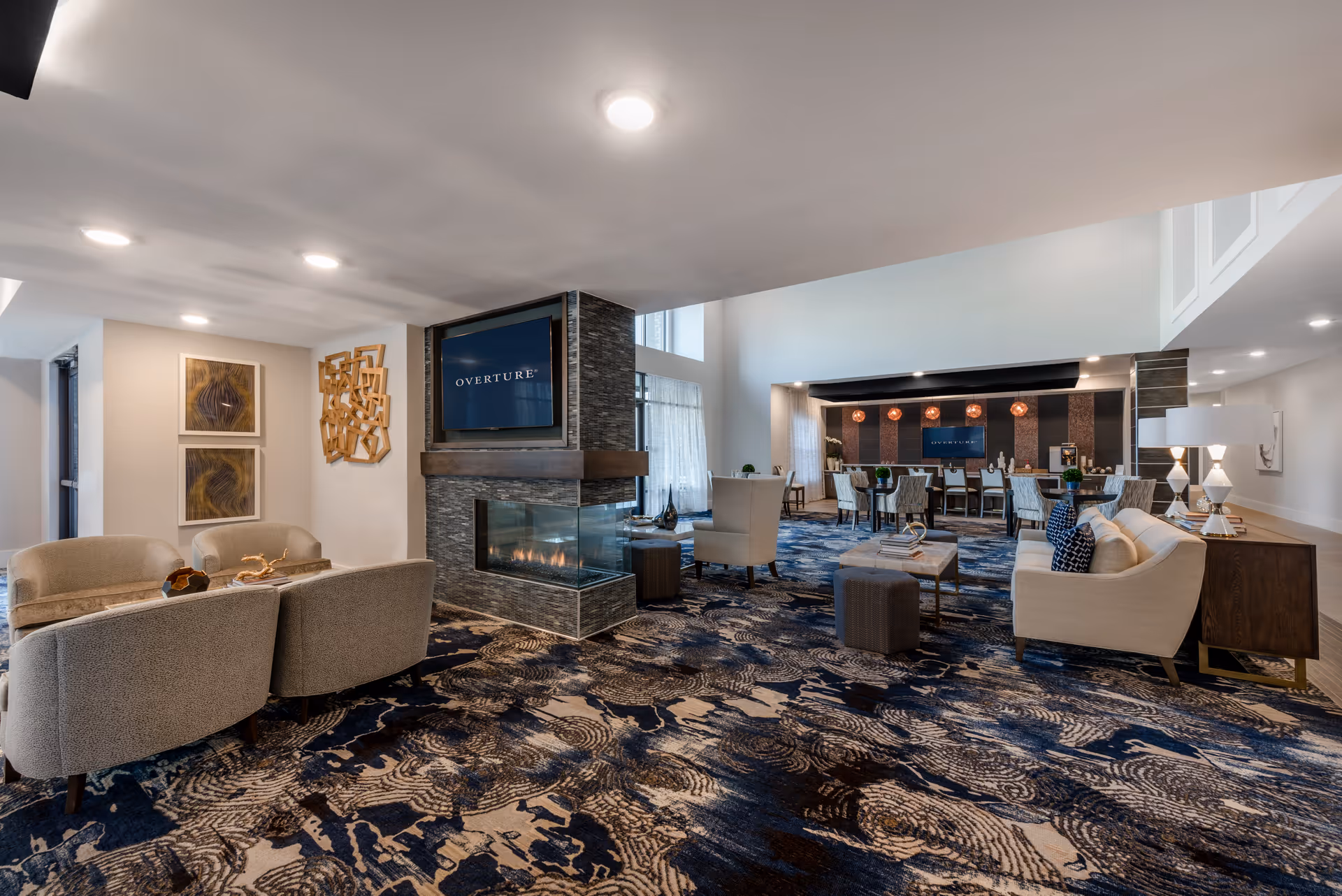 A spacious and modern living room area in a senior living facility featuring a patterned carpet, multiple seating arrangements including armchairs and sofas, a central fireplace with a mounted TV displaying 'OVERTURE', decorative wall art, and a dining area with tables and chairs in the background.