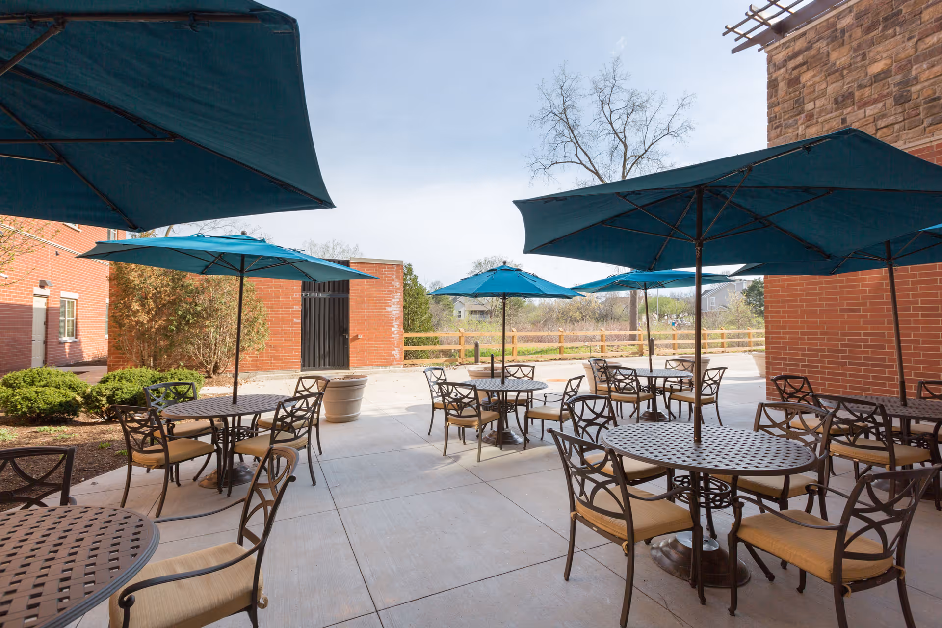 Outdoor patio area with multiple round metal tables and chairs, each table shaded by a large blue umbrella. The patio is surrounded by brick walls and some greenery, with a clear sky overhead.