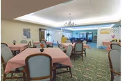 A spacious dining room with multiple tables covered in pink tablecloths, surrounded by cushioned chairs. The room features a patterned green carpet, a chandelier hanging from the ceiling, and large windows allowing natural light. There are decorative plants and artwork on the walls, and an open doorway leading to another room.