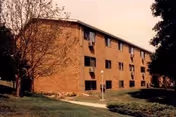 Exterior brick apartment building with multiple stories, trees, and a walkway along a grassy lawn.