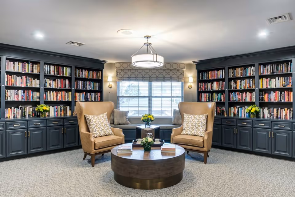 A cozy library room with two tan leather armchairs, each with a decorative pillow, positioned around a round wooden coffee table with books and a small flower arrangement. The room features built-in dark gray bookshelves filled with books and decorated with yellow flower vases. A window with patterned Roman shades is centered between the bookshelves, with a cushioned bench underneath. The ceiling has recessed lighting and a modern circular chandelier.