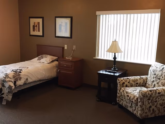 A simple bedroom with a single bed covered in white bedding with floral patterns, a wooden nightstand with a telephone, a window with vertical blinds, a table lamp on a small dark wooden side table, and a patterned armchair.