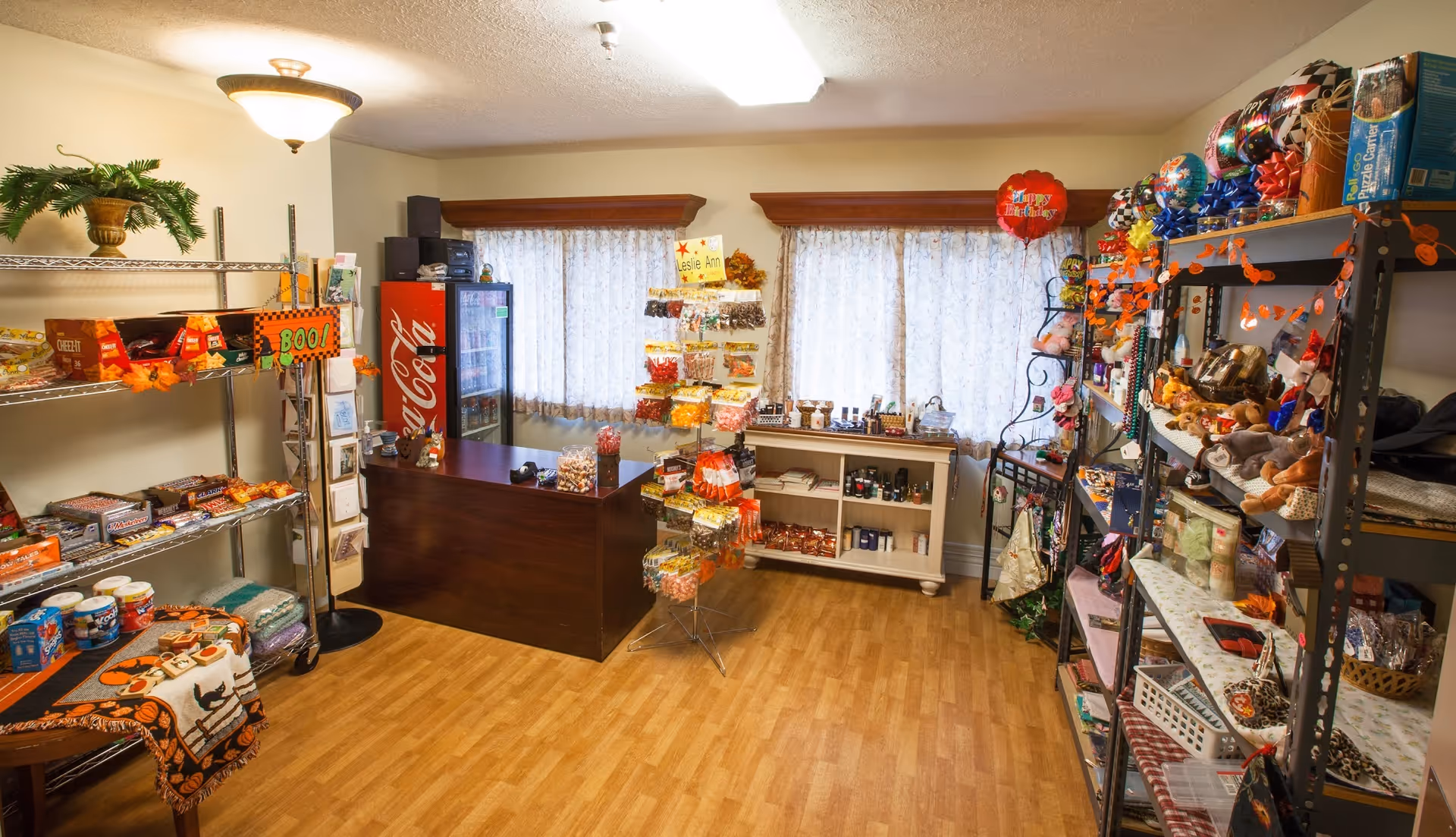 Interior view of a small gift shop or snack area in a senior living facility with shelves stocked with snacks, candies, and small gift items. There is a wooden counter with a Coca-Cola refrigerator behind it, two windows with lace curtains, and various decorations including a 'Boo!' sign and balloons.