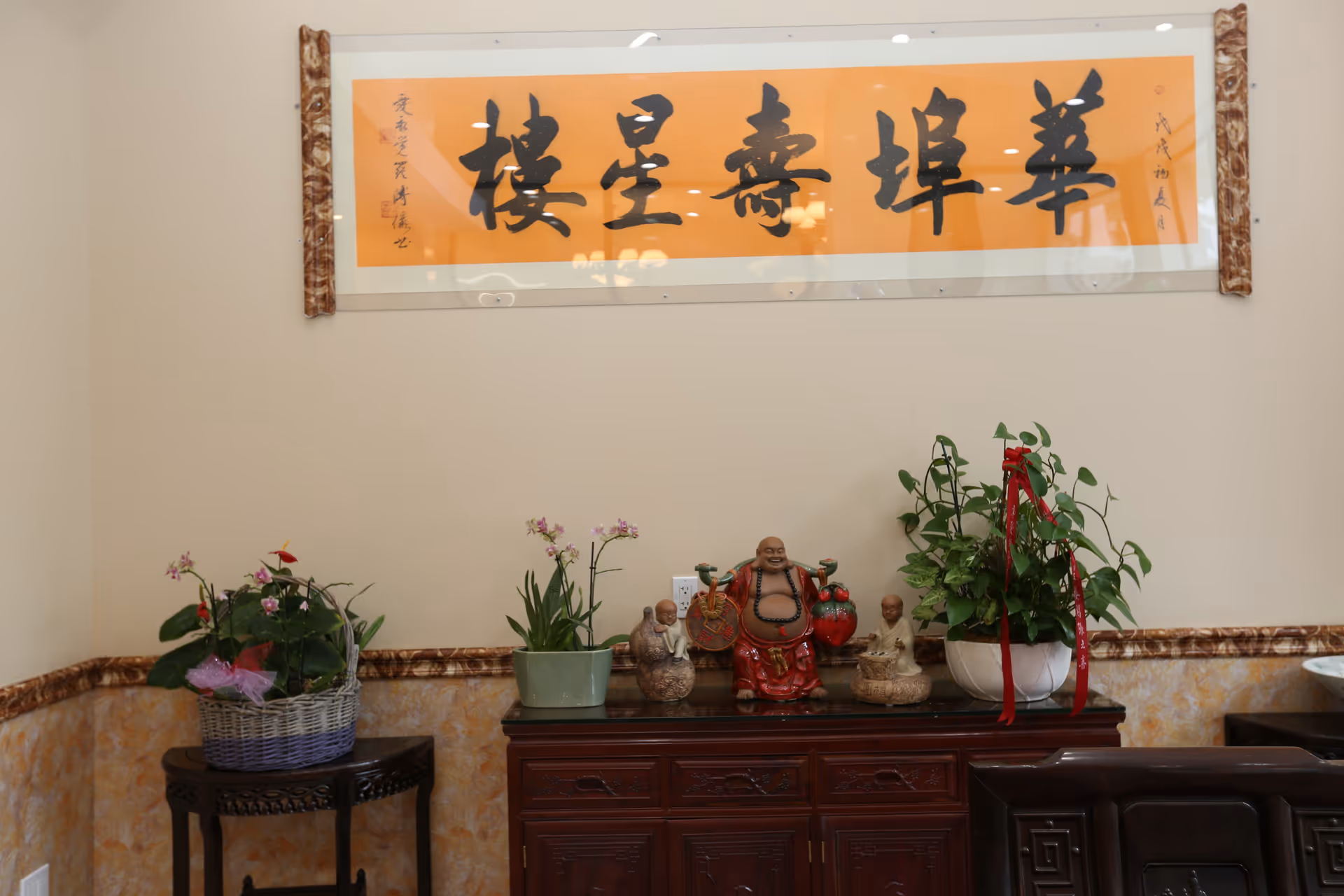 An interior corner of a room with a wooden cabinet topped with decorative statues including a laughing Buddha and two smaller figurines, flanked by potted plants. Above the cabinet is a large framed orange scroll with black Chinese calligraphy. The walls are light-colored with a decorative trim.