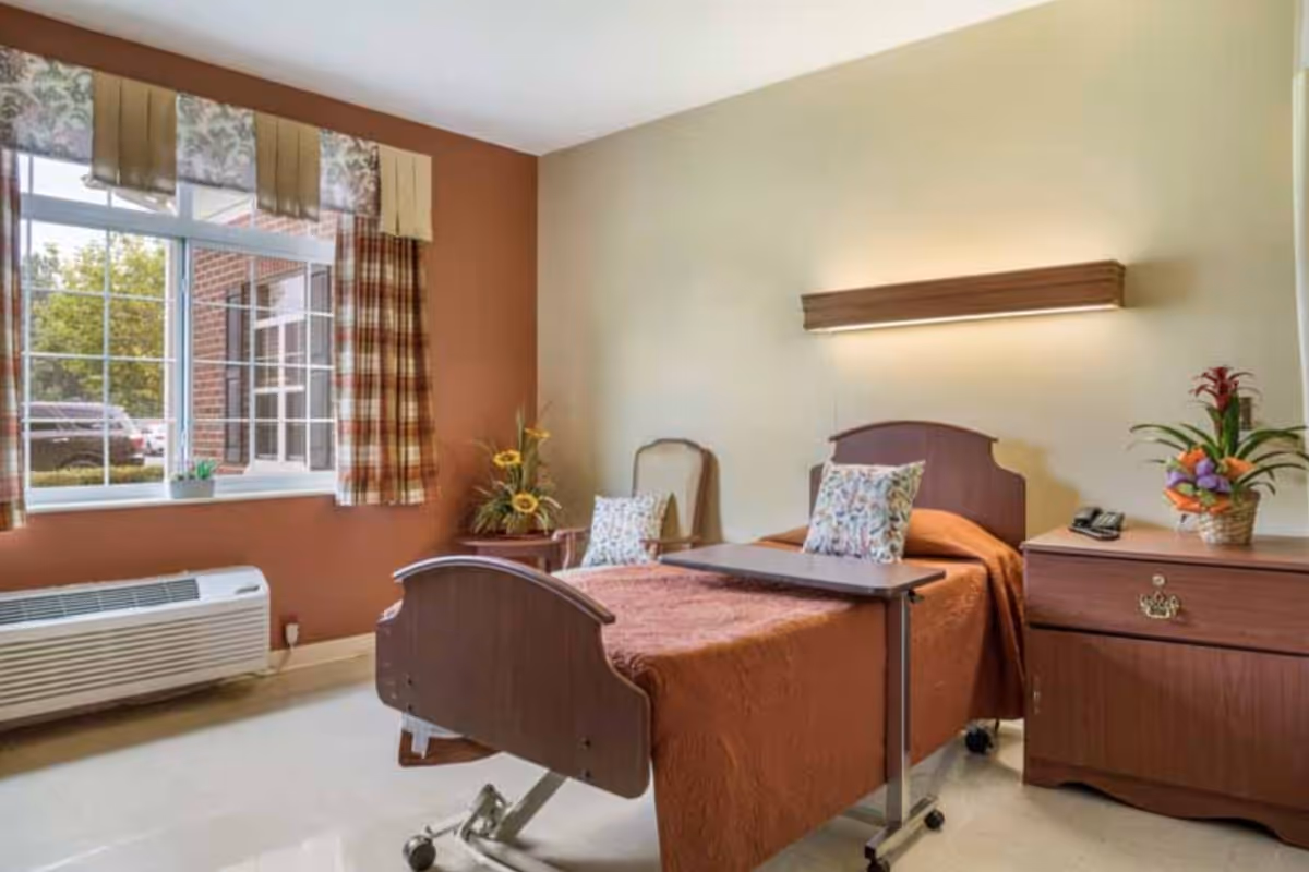 A single hospital-style bed with a brown bedspread and floral pillows in a well-lit room. The room has a large window with plaid curtains, a wooden nightstand with a telephone and a flower arrangement, a chair with a floral cushion, and a wall-mounted light above the bed. There is also a small overbed table on wheels and a heating/cooling unit below the window.