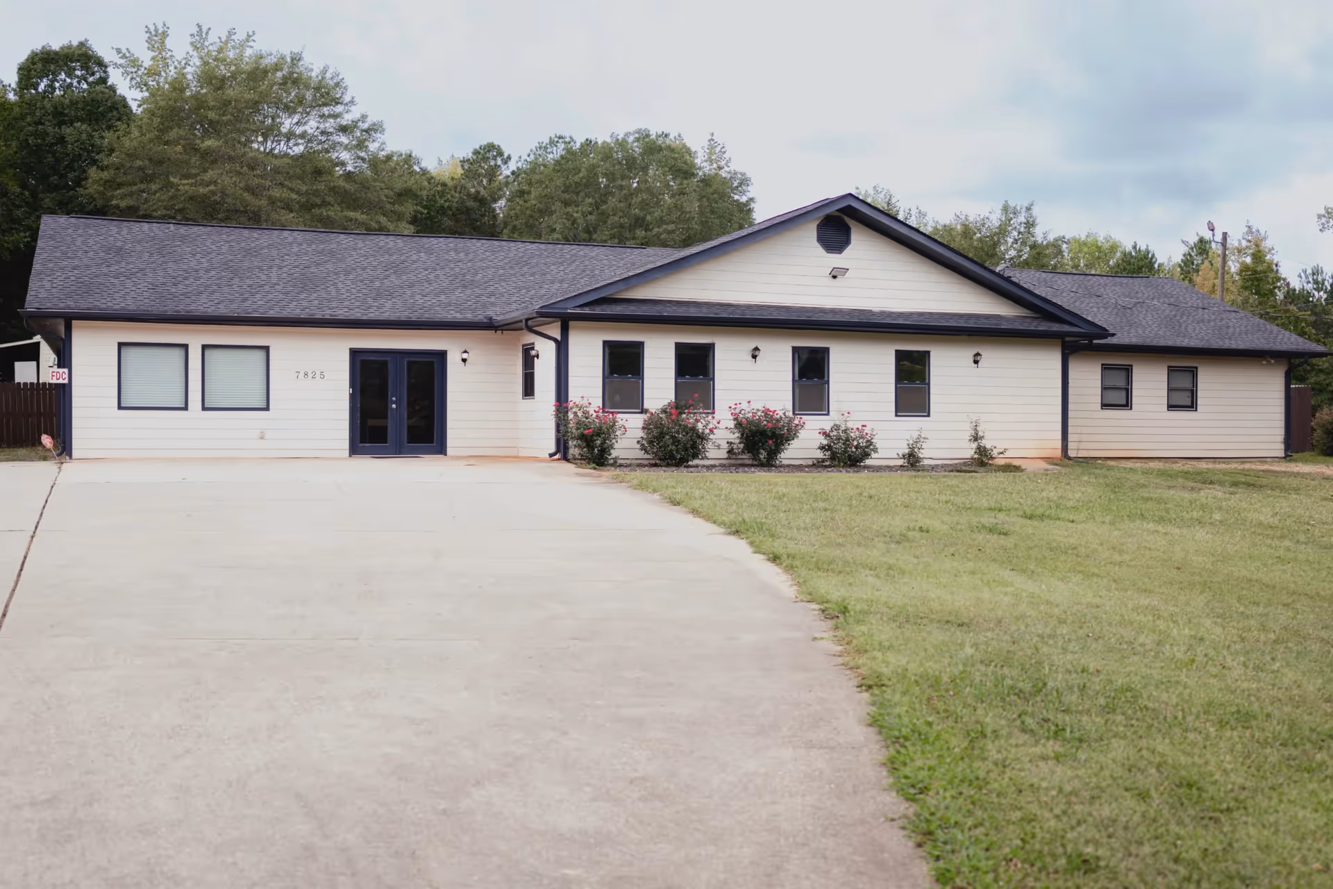 Single-story building with beige siding and a dark roof, surrounded by grass and trees. The building has several windows and a double-door entrance with the number 7825 visible on the front. There is a concrete driveway leading up to the entrance.
