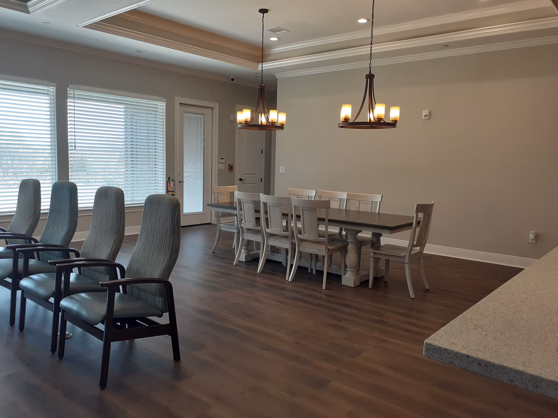 A spacious dining area with a long rectangular table surrounded by white wooden chairs. There are two modern chandeliers hanging from the ceiling above the table. To the left, there are several high-backed chairs with armrests arranged in a row near large windows with blinds. The floor is wooden, and the walls are painted light beige. A door with glass panels is visible in the background.