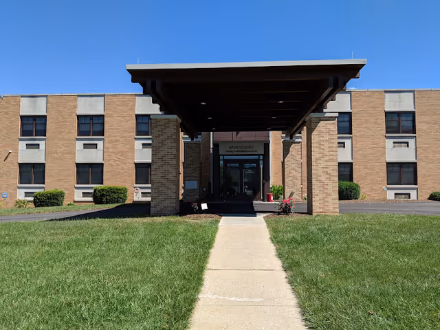 Front entrance of Misericordia Nursing & Rehabilitation Center showing a brick facade, covered portico, and a walkway through a grassy lawn under a clear blue sky.