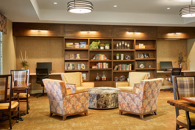 Cozy common room with floral and upholstered armchairs arranged around a round ottoman facing a built-in bookshelf and work desks.