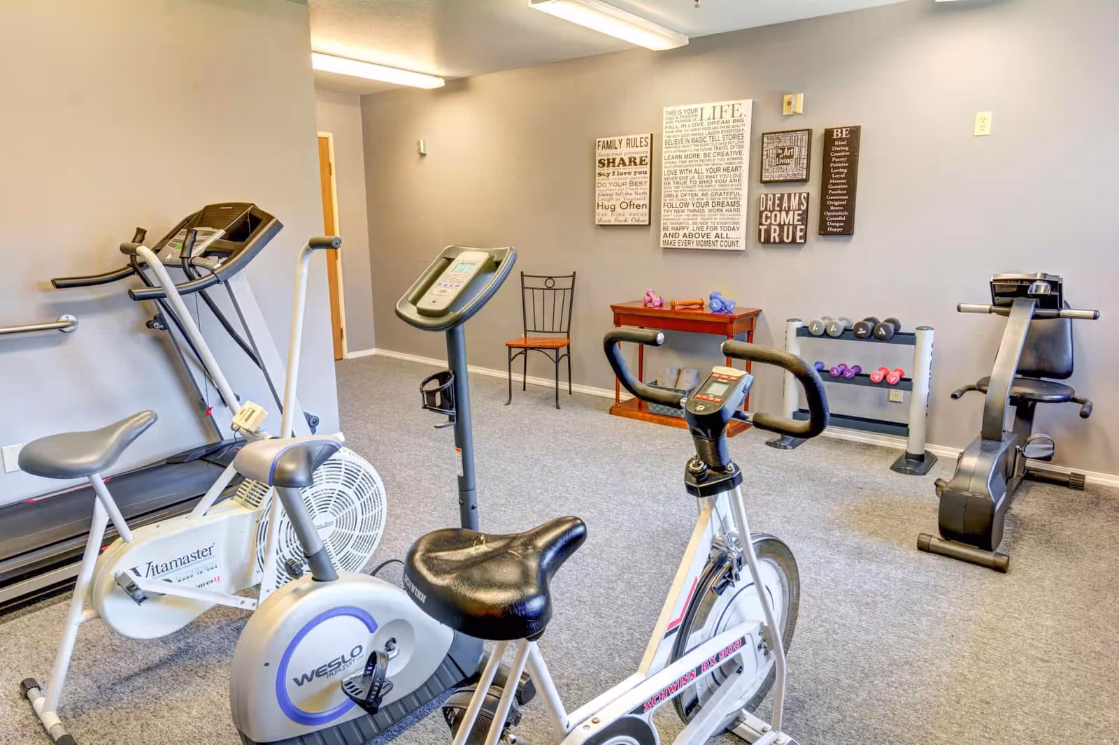 A small fitness room with exercise equipment including a treadmill, two stationary bikes, a rowing machine, and a rack of dumbbells. The walls are decorated with motivational and family-themed wall art, and there is a chair and a small wooden table with additional weights on it.