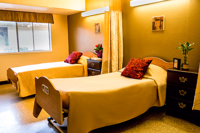 A room with two single beds, each covered with a beige blanket and a red decorative pillow. Between the beds are two dark wooden nightstands, one with a vase of red flowers and the other with a small plant and framed photo. The room has a window with a brown valance and a beige curtain partially drawn between the beds. The walls are painted a warm beige color and there is a small framed artwork on the wall.