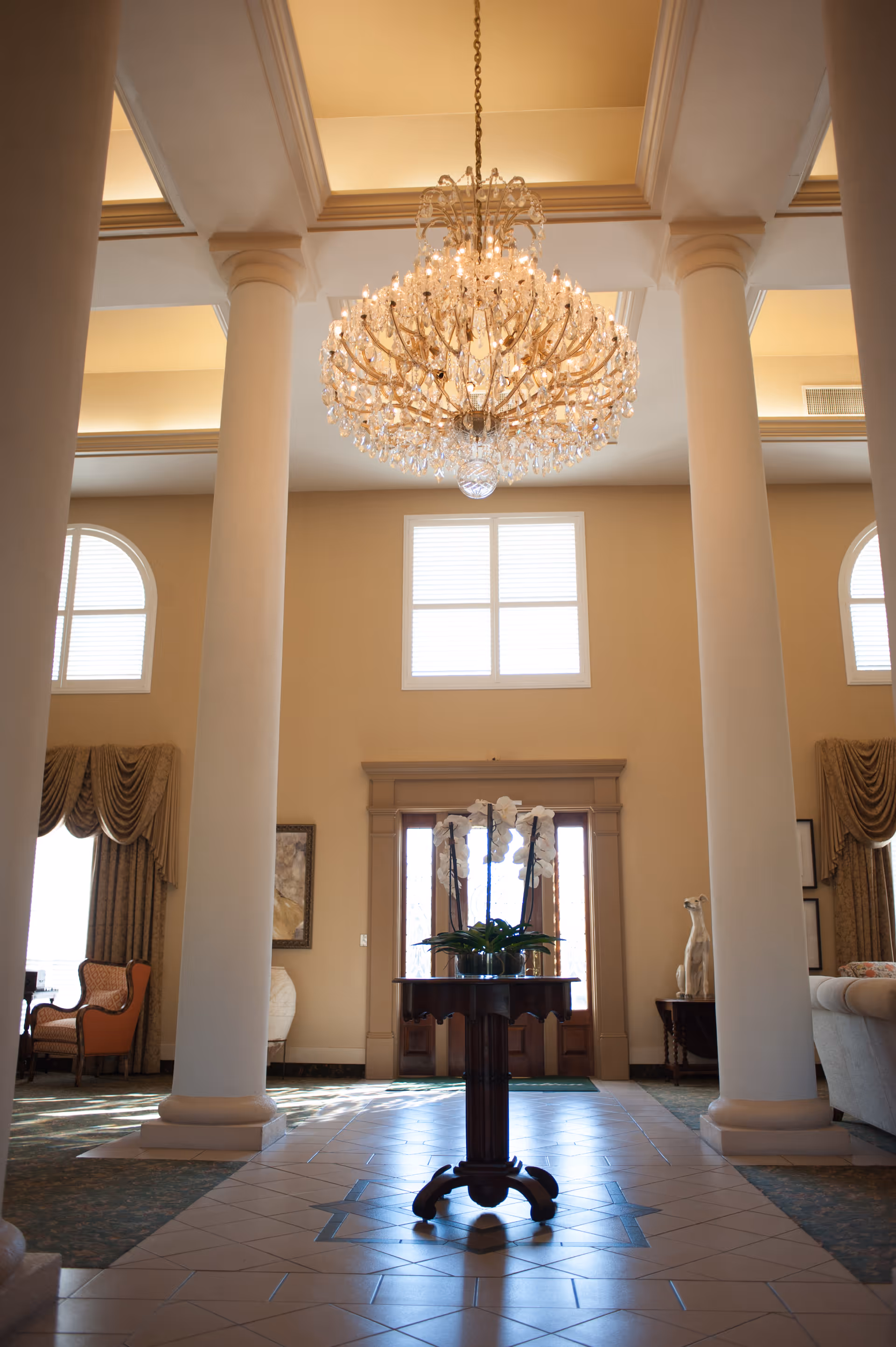 Interior view of a spacious and elegant living area with tall white columns, a large crystal chandelier hanging from the ceiling, a round wooden table with a white orchid plant in the center, and large windows letting in natural light. The room features beige walls, patterned curtains, and comfortable seating including an armchair and a sofa.