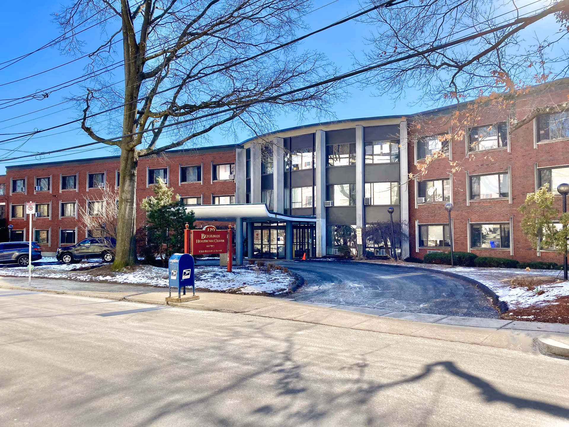 Exterior view of Edgar P. Benjamin Healthcare Center, a three-story brick building with multiple windows. There is a driveway leading to the main entrance, a red sign with the facility name, a blue USPS mailbox, and some parked cars. Trees with bare branches and patches of snow are visible around the building under a clear blue sky.