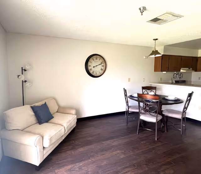 Open living area with a beige loveseat, round dining table and chairs, wall clock, and a view into the kitchen.