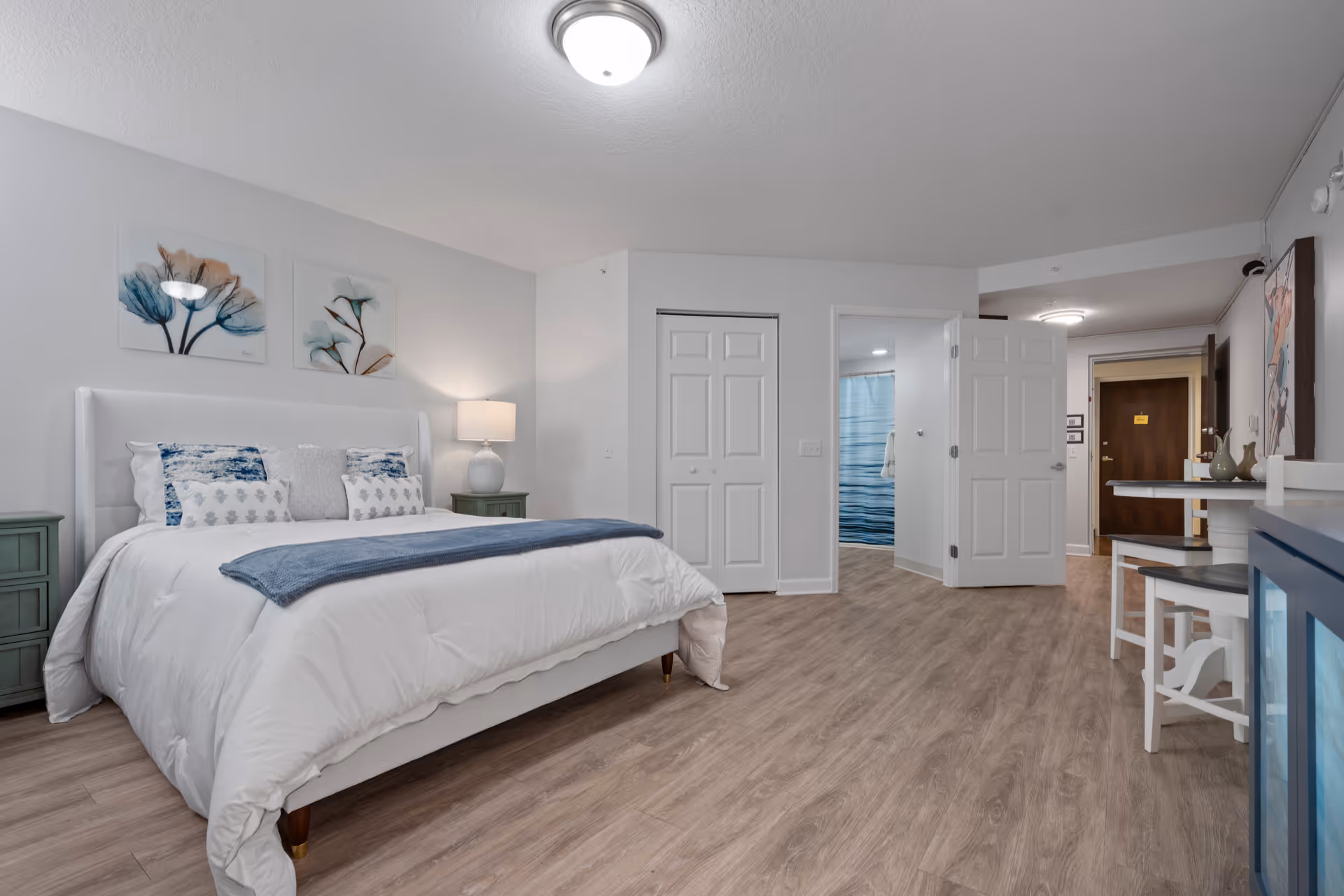 A spacious bedroom with a large white bed adorned with multiple pillows and a blue throw blanket. There are two green nightstands on either side of the bed, one with a lamp. The room has light wood flooring and white walls decorated with two floral paintings above the bed. An open door reveals a bathroom with a blue shower curtain, and another door leads to a hallway with a dark wooden door at the end. A small dining area with a white table and chairs is visible on the right side.