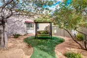 Outdoor courtyard area at Life Spire Assisted Living Rio Rancho featuring a green artificial turf pathway leading to a covered swing bench surrounded by trees and shrubs, with the building exterior visible in the background.