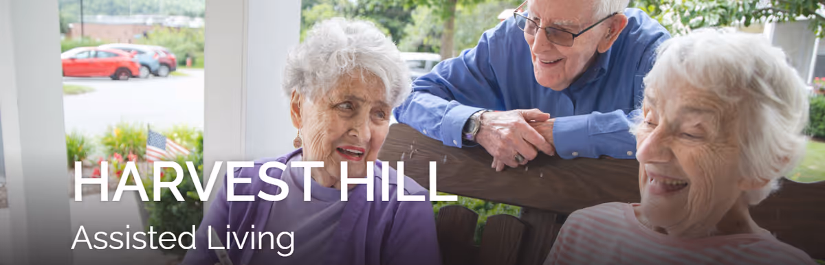 Three elderly people sitting and smiling together on a wooden bench outdoors with greenery and parked cars in the background. The text 'HARVEST HILL Assisted Living' is overlaid on the image.
