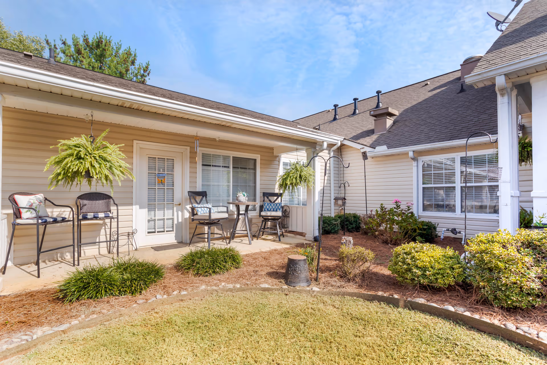 Outdoor patio area at Brookdale Sandy Springs with beige siding walls, two black chairs with cushions, a small round table, hanging green ferns, and landscaped garden beds with shrubs and grass under a blue sky.