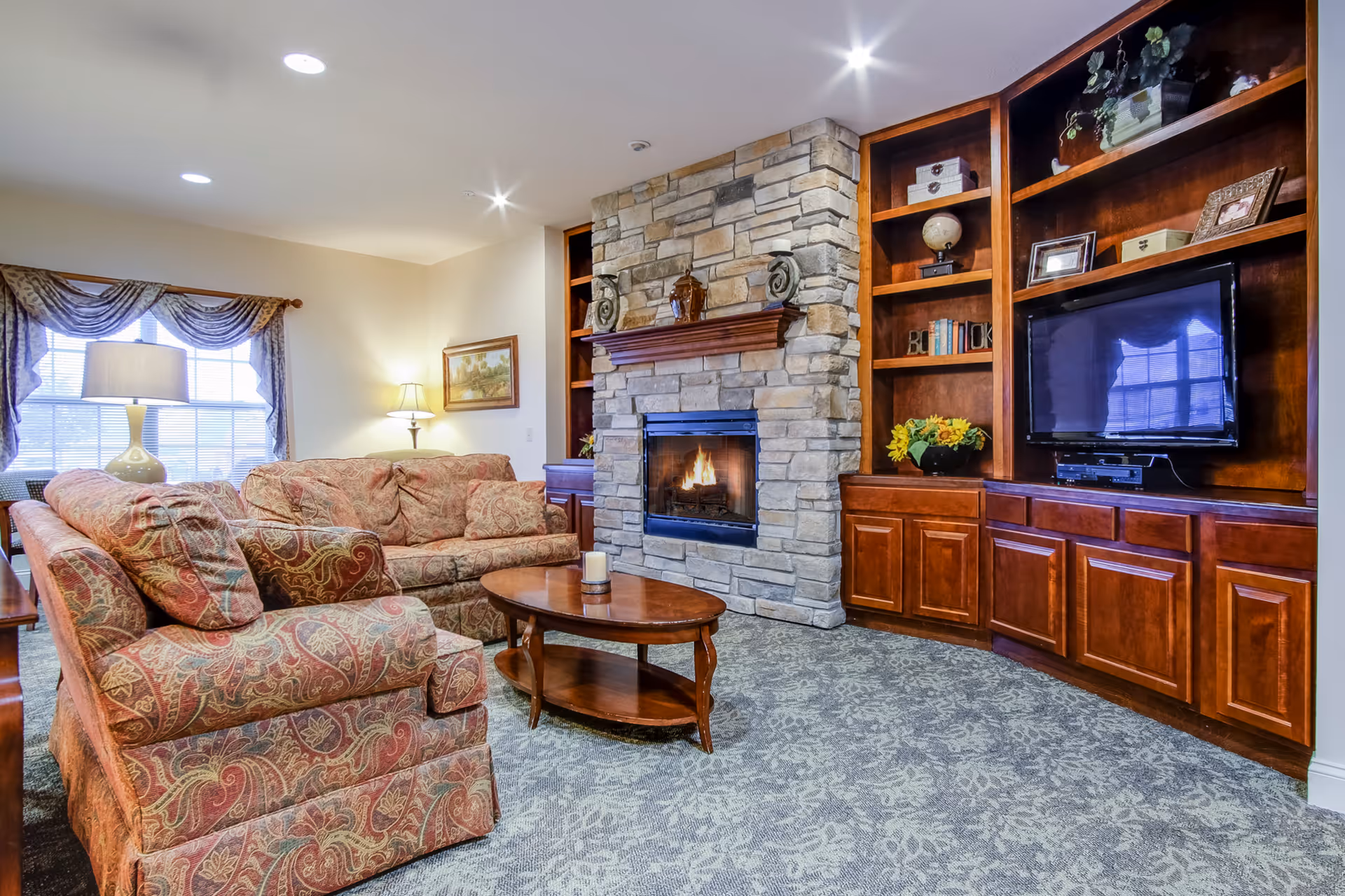 Cozy living room with patterned sofas, a central coffee table, stone fireplace, and built-in wooden shelving with a TV.