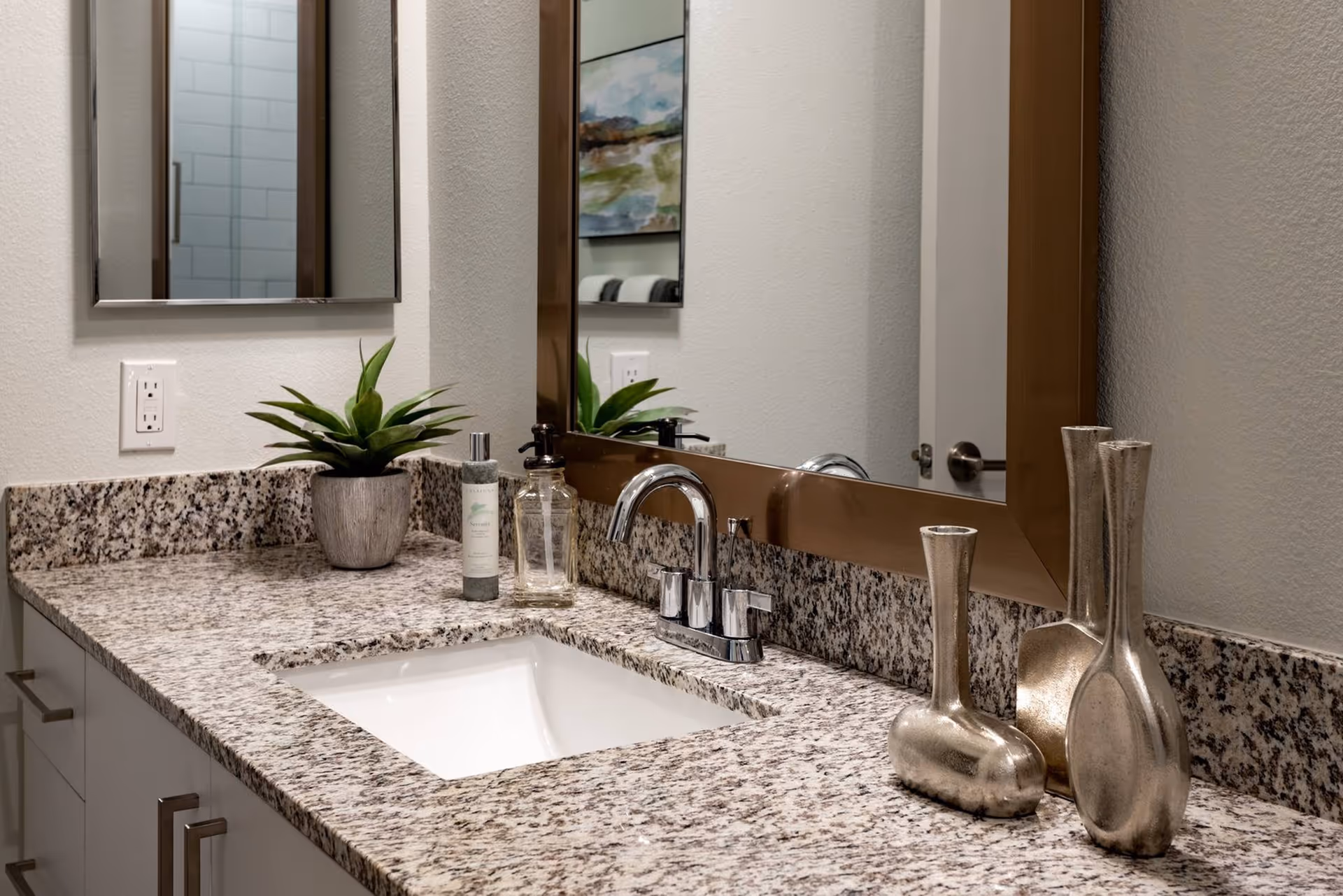 Granite countertop bathroom vanity with undermount sink, chrome faucet, large mirror, potted plant, soap dispensers and decorative vases.