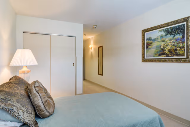 A bedroom in a senior living facility featuring a bed with decorative pillows and a light blue bedspread. A bedside lamp with a white shade is on a nightstand next to the bed. There is a sliding closet door on the left wall and a framed painting of a garden scene on the right wall. A hallway with a wall-mounted mirror and a light fixture is visible in the background.
