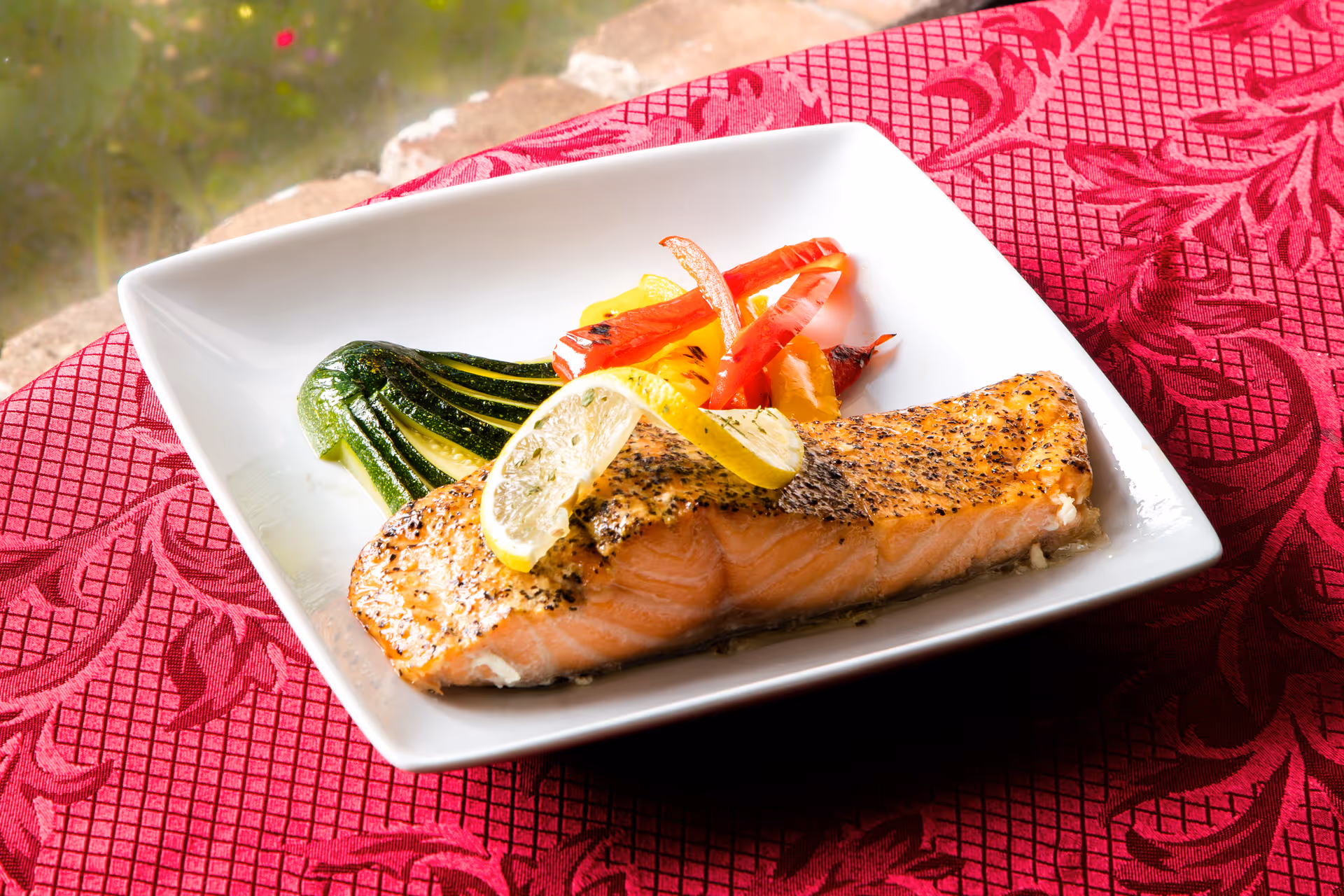 A grilled salmon fillet topped with a lemon slice served with roasted vegetables on a white square plate on a red patterned tablecloth.
