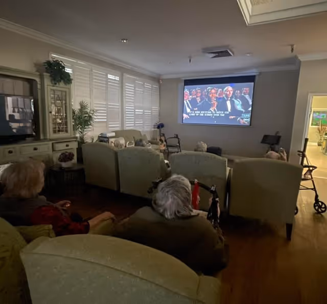 A group of elderly people seated in a dimly lit living room watching a movie projected on a screen. The room has comfortable armchairs, a TV on a cabinet, plants, and window shutters. Some walkers are visible near the chairs.