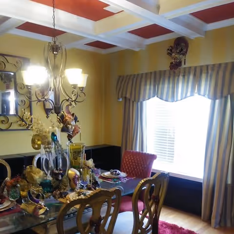 A warmly lit dining room with a glass-top table set with decorative items and place settings. The room features a chandelier with multiple lights hanging above the table, a large window with striped curtains, and a red upholstered chair. The walls are painted yellow with a white and red coffered ceiling.