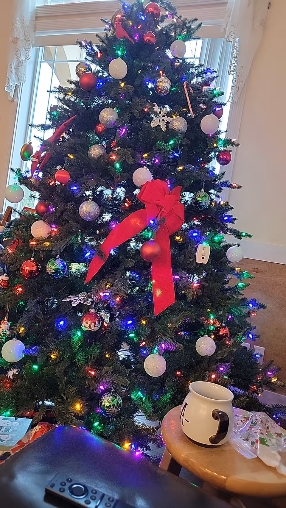 A decorated Christmas tree with multicolored lights, red and white ornaments, and a large red bow in front of a window with white curtains. In the foreground, there is a wooden table with a white mug and a black remote control on a dark surface.