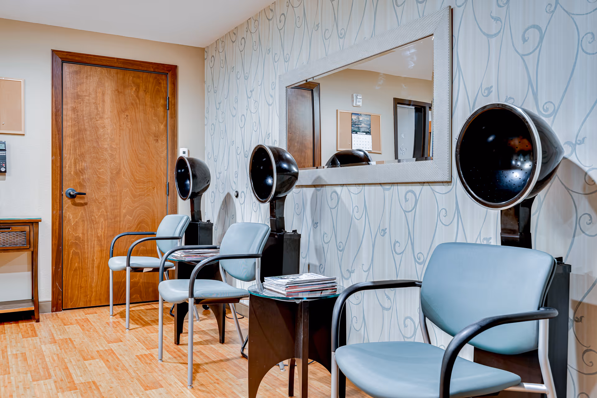 A salon waiting area with three hooded hair dryers, blue chairs, a large wall mirror and a small table with magazines.