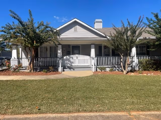 Front exterior view of a single-story senior living facility with a white porch railing, two trees in the front yard, and a well-maintained lawn under a clear blue sky.