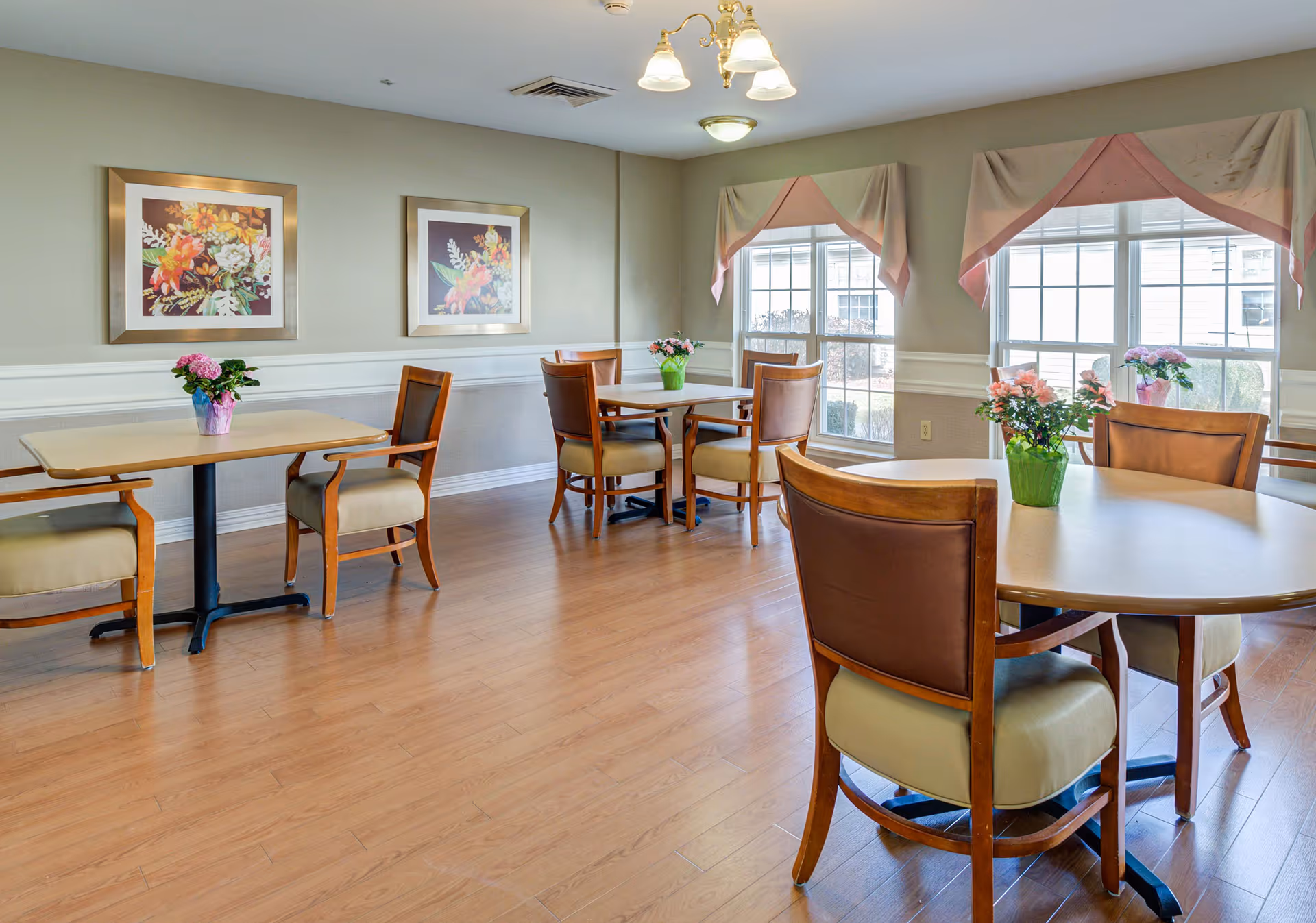 Bright communal dining room with round and rectangular tables, wooden chairs, floral centerpieces, framed artwork, and large windows.
