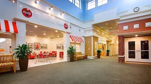 Interior view of a senior living facility hallway with a dining area named 'Paula's Street Shoppe' on the left, featuring red and white striped awnings, red chairs, and tables. The hallway has beige carpeting, potted plants, benches, and a high ceiling with windows allowing natural light.