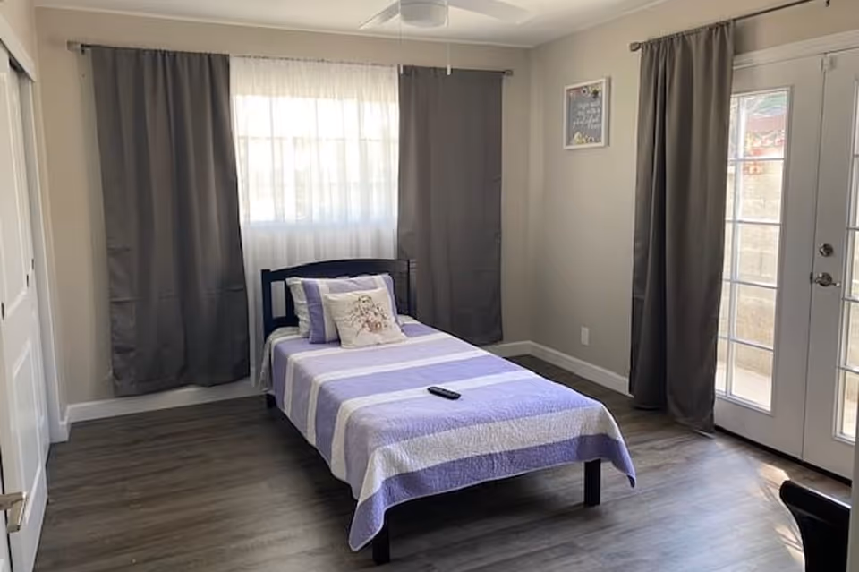 A simple bedroom with a single bed covered in a purple and white striped quilt, a decorative pillow, and a remote control on the bed. The room has gray curtains on the windows and glass doors, light-colored walls, and wood flooring.