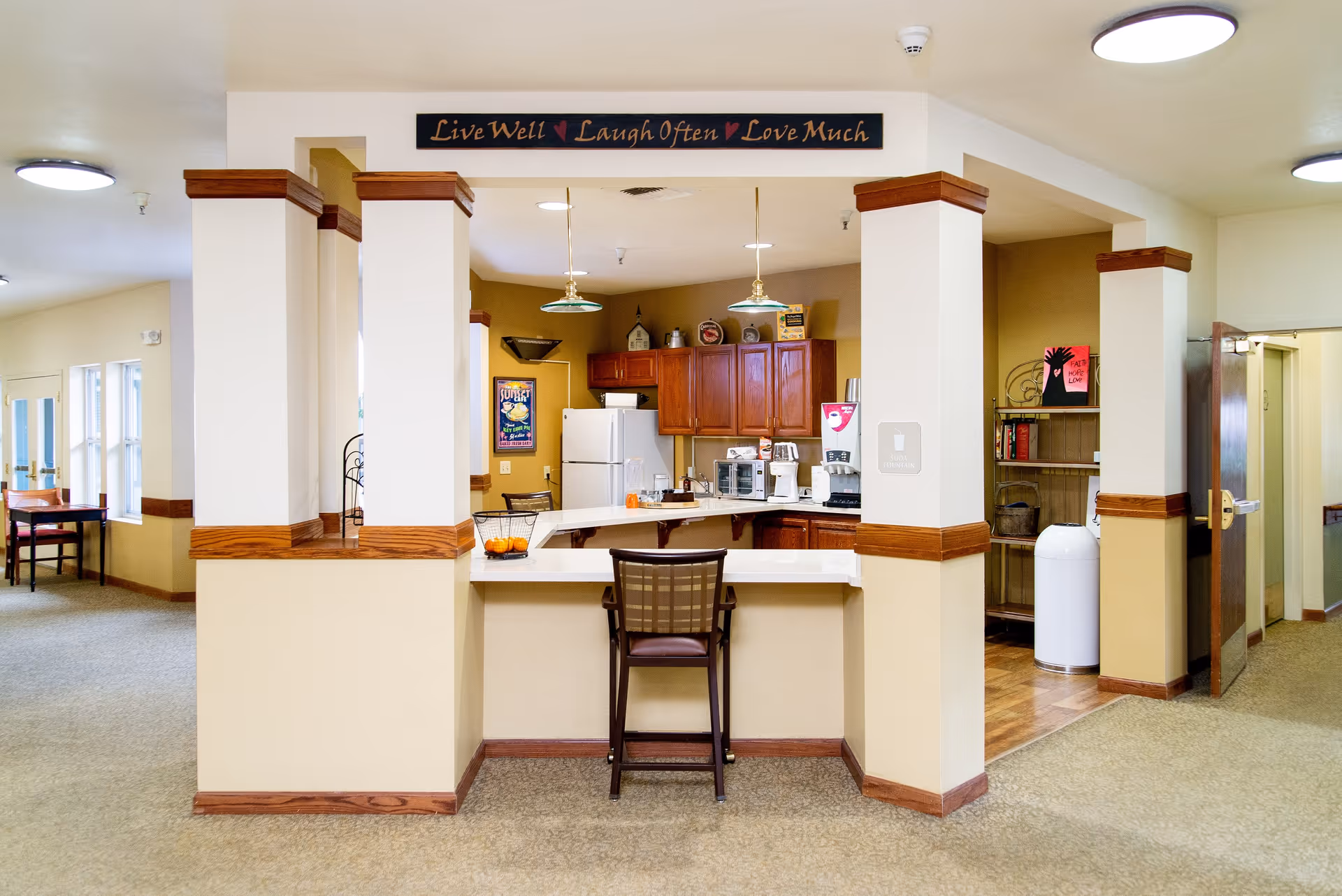 Interior view of a senior living facility kitchen area with a counter and a single chair in front. The kitchen has wooden cabinets, a refrigerator, a microwave, a coffee maker, and a soda fountain machine. Above the kitchen area is a decorative sign that reads 'Live Well Laugh Often Love Much'. The surrounding area has beige walls with wooden trim and carpeted flooring.
