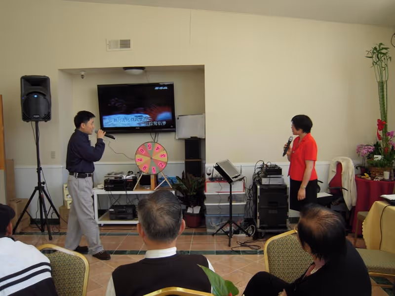 Two people holding microphones appear to be singing karaoke in a room with a television screen displaying Chinese text. Several seated people watch them. The room has audio equipment, a speaker, a colorful spinning wheel, and some plants and furniture.