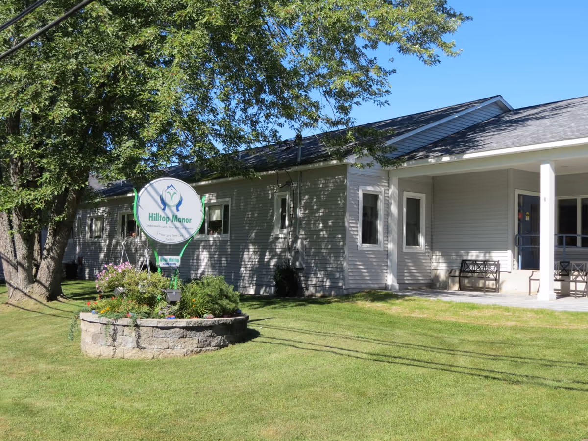 Exterior view of Hilltop Manor, a single-story building with light gray siding and a covered porch area with benches. There is a large tree and a circular stone planter with flowers and greenery in front of the building under a clear blue sky.